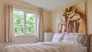 Un dormitorio con una cama, una ventana con cortinas, una mesita de noche y un cuadro en la pared.