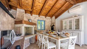 A kitchen with a brick oven, wooden ceiling beams, and a dining area with white chairs and a table set for a meal. - Villa Milo Mali Vareski With Pool (Marcana)