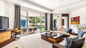 A modern living room with a large TV, comfortable seating, a glass coffee table, and a view of a pool and garden.