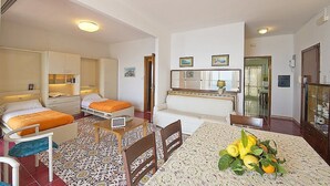 Habitación de hotel con dos camas, una mesa de comedor con un centro de frutas, un televisor pequeño y una kitchenette.