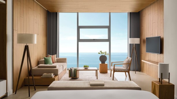 Una habitación de hotel moderna con una cama grande, un sofá, una silla, una mesita de café y un televisor. La habitación tiene paredes de madera y un ventanal con vistas al mar.