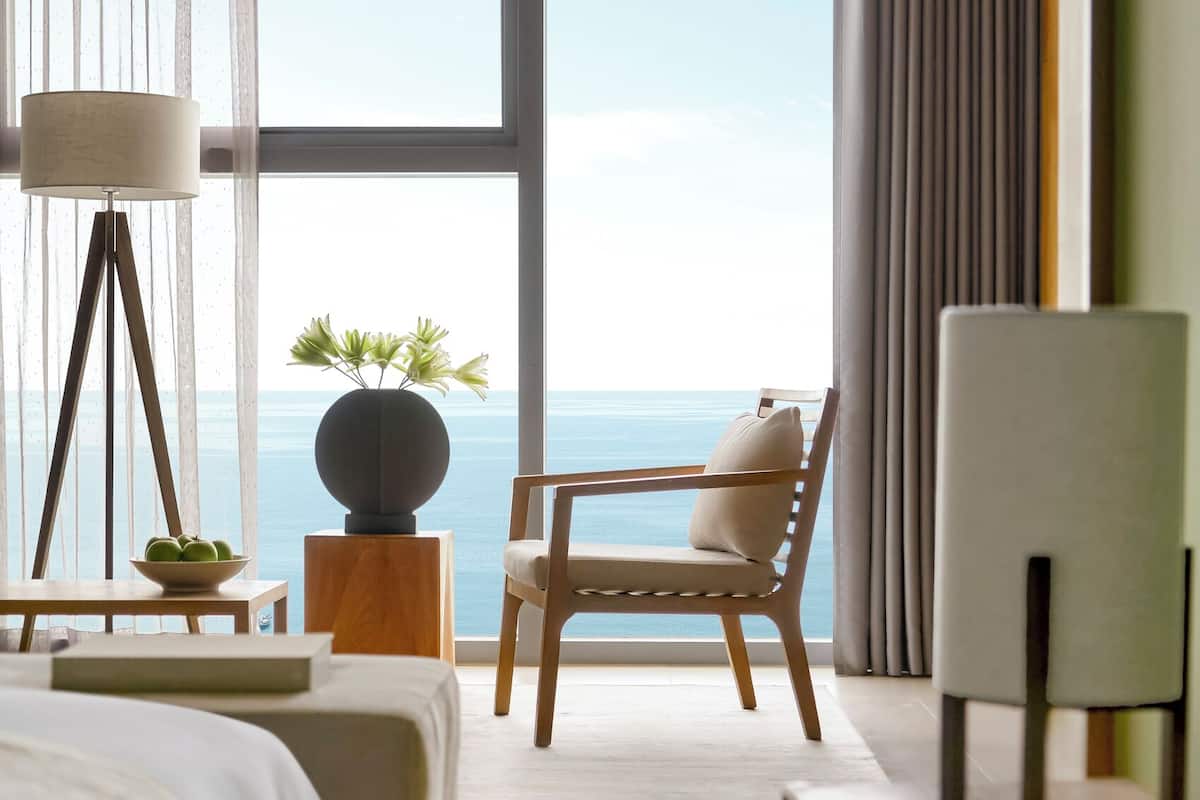 A modern hotel room with a large window, a bed, a chair, a lamp, and a small table with a plant.