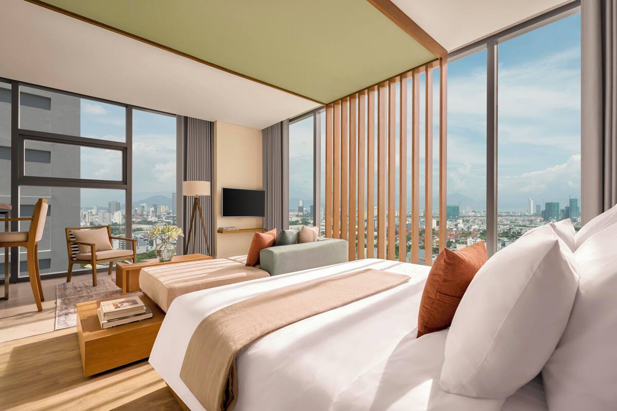 A modern hotel room with a large bed, a sofa, a desk, and a chair. There is a large window with a city view.