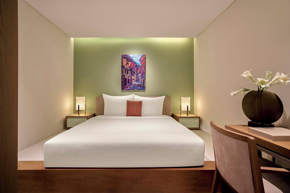 A modern hotel room with a large bed, bedside tables with lamps, a painting on the wall, and a vase with flowers on a side table.