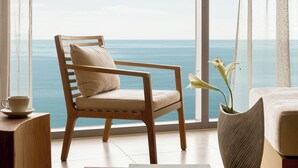 Una habitación con un ventanal que ofrece vistas al mar, una silla de madera, un jarrón con flores y un libro sobre una mesa.