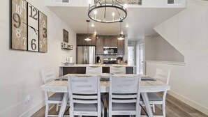 A dining area with a wooden table and white chairs, a chandelier, and a kitchen in the background.