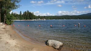 A lakeside beach with clear water, a large rock in the foreground, and a forested shoreline.
