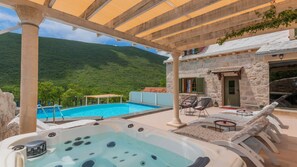 Un espace piscine doté d’un jacuzzi, de chaises longues et d’un bâtiment en pierre offrant une vue sur des collines verdoyantes.