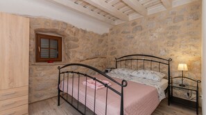 Une chambre avec un mur en pierre, une armoire en bois, un lit avec une literie rose, une fenêtre avec des volets et une lampe de chevet.