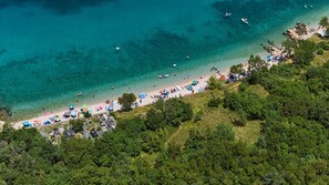 An aerial view of a beach with clear turquoise water, a sandy shore, and dense green foliage.