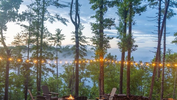 An outdoor seating area with Adirondack chairs, a fire pit, and string lights against a backdrop of tall trees and a view of the water. - Private 6-Acre Retreat | Views, Hot Tub, Fire Pit (Ellijay)