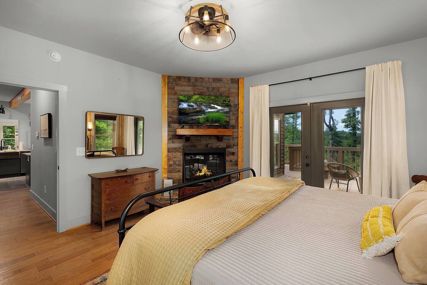 A bedroom with a bed, a dresser, a mirror, a fireplace, and a balcony with a view of the outdoors.