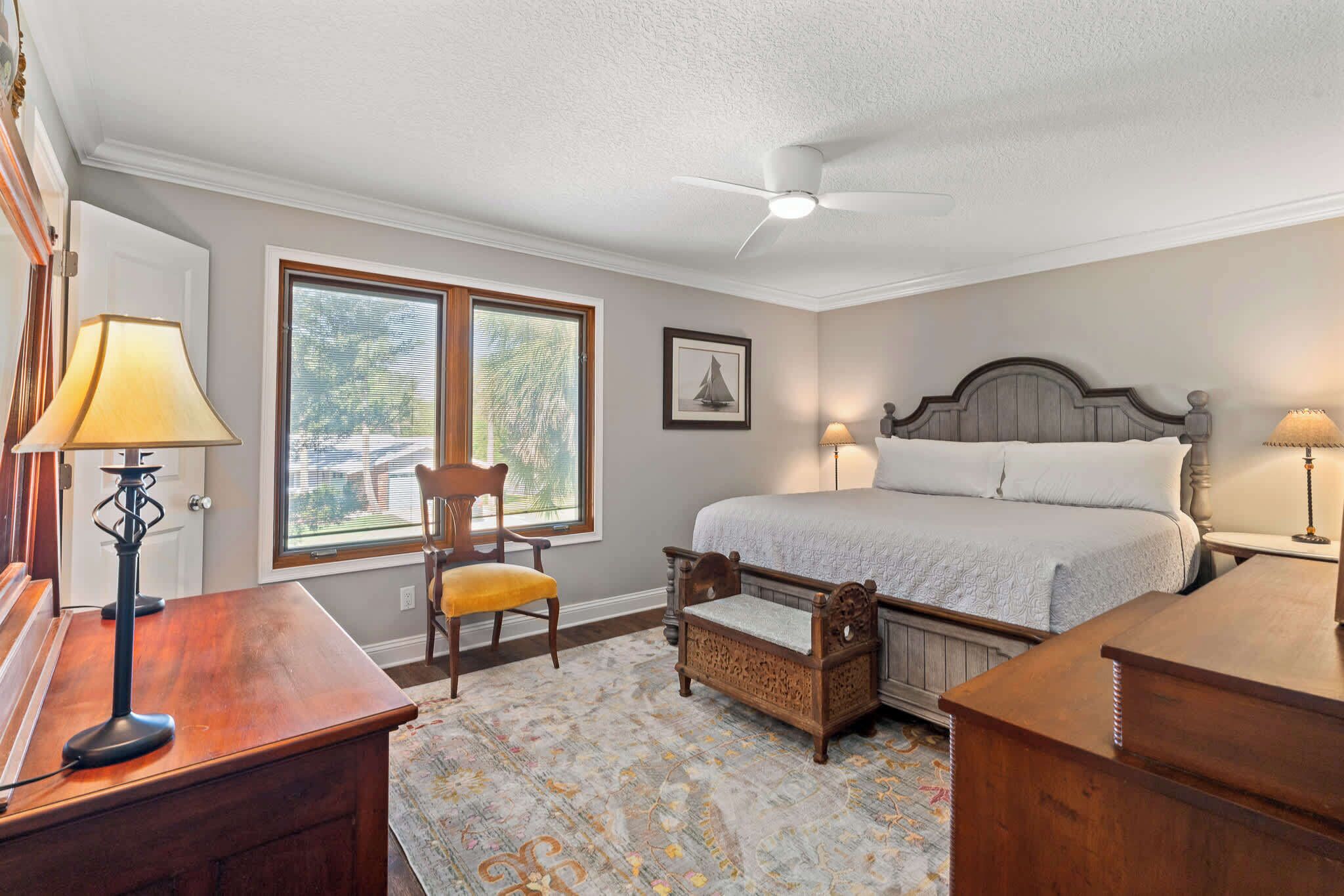A bedroom with a bed, a desk, a chair, a desk lamp, a standing lamp, a ceiling fan, and a window with blinds.
