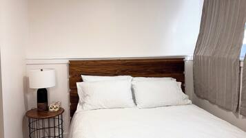 Una cama bien hecha con sábanas blancas, una mesita de noche redonda con una lámpara y un cabecero de listones de madera verticales.