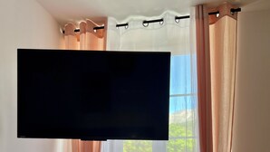 A bedroom with a large flat-screen TV mounted on the wall, a wooden dresser, and a window with sheer curtains. - Villa Anita -town center -private parking- wifi. (Barcelonnette)