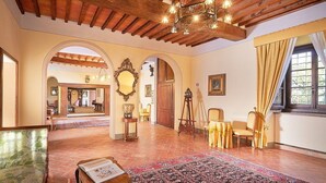 A spacious room with a high ceiling, wooden beams, and a large mirror. There are multiple seating areas with chairs and a sofa. The room has a tiled floor and is decorated with framed pictures and a chandelier. - Villa Rama: A splendid seventeenth-century two-story historical villa surrounded by meadows and by green hills, with Free WI-FI. (Gambassi Terme)