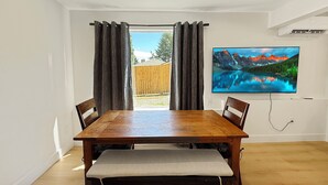 Een eetruimte met een houten tafel en stoelen, een bank, een grote flatscreen televisie aan de muur, en uitzicht op een houten omheining en groen buiten.