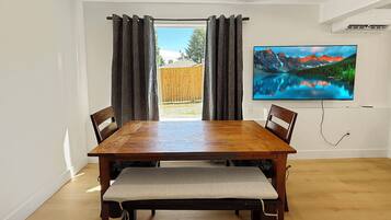 A dining area with a wooden table and chairs, a bench, a large flat-screen TV on the wall, and a view of a wooden fence and greenery outside.