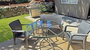 Een terras met een glazen tafel, omringd door rieten stoelen, een stenen muur en een houten omheining op de achtergrond.