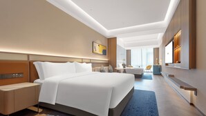 Moderni hotellihuone, jossa on suuri sänky, yöpöydät, työpöytä sekä oleskelualue, jossa on tuoli ja pieni pöytä.