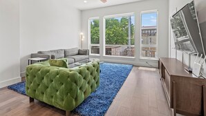Een moderne woonkamer met een grijze bank, een groen gestoffeerde fauteuil, een houten salontafel en een blauw vloerkleed. Er zijn grote ramen met uitzicht op bomen en gebouwen.