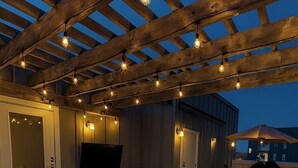 Een buitenterras met een houten pergola, een zithoek en een flatscreen televisie aan een muur.