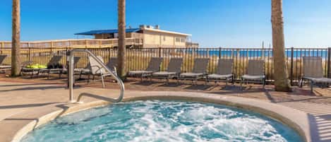 A hot tub area with lounge chairs and a pool, surrounded by a fence and palm trees.