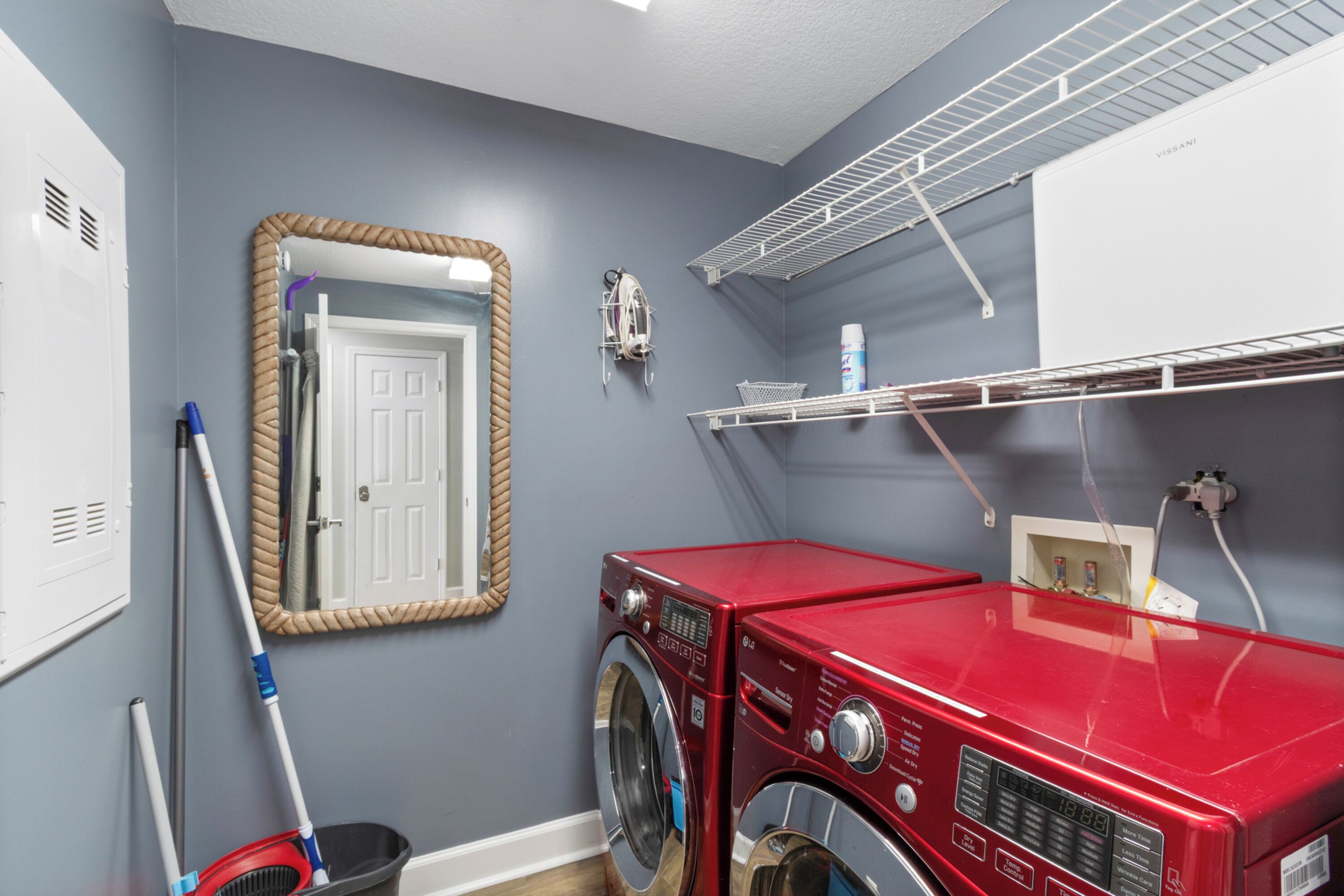 A laundry room with a red washing machine, a mop, a broom, and a mirror on the wall.