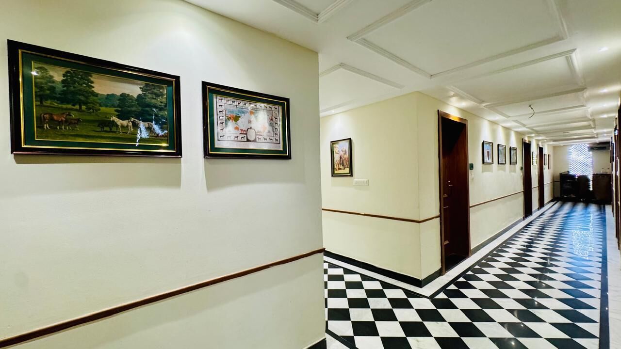 A hotel corridor with framed artwork on the walls, a checkered floor, and doors leading to rooms.
