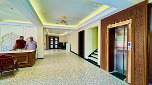 Vestíbulo de hotel con mostrador de recepción, alfombra estampada, ventilador de techo y ascensor.