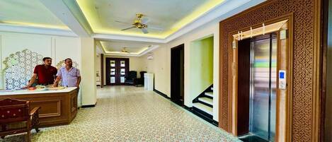A hotel lobby with a reception desk, a patterned carpet, a ceiling fan, and an elevator.