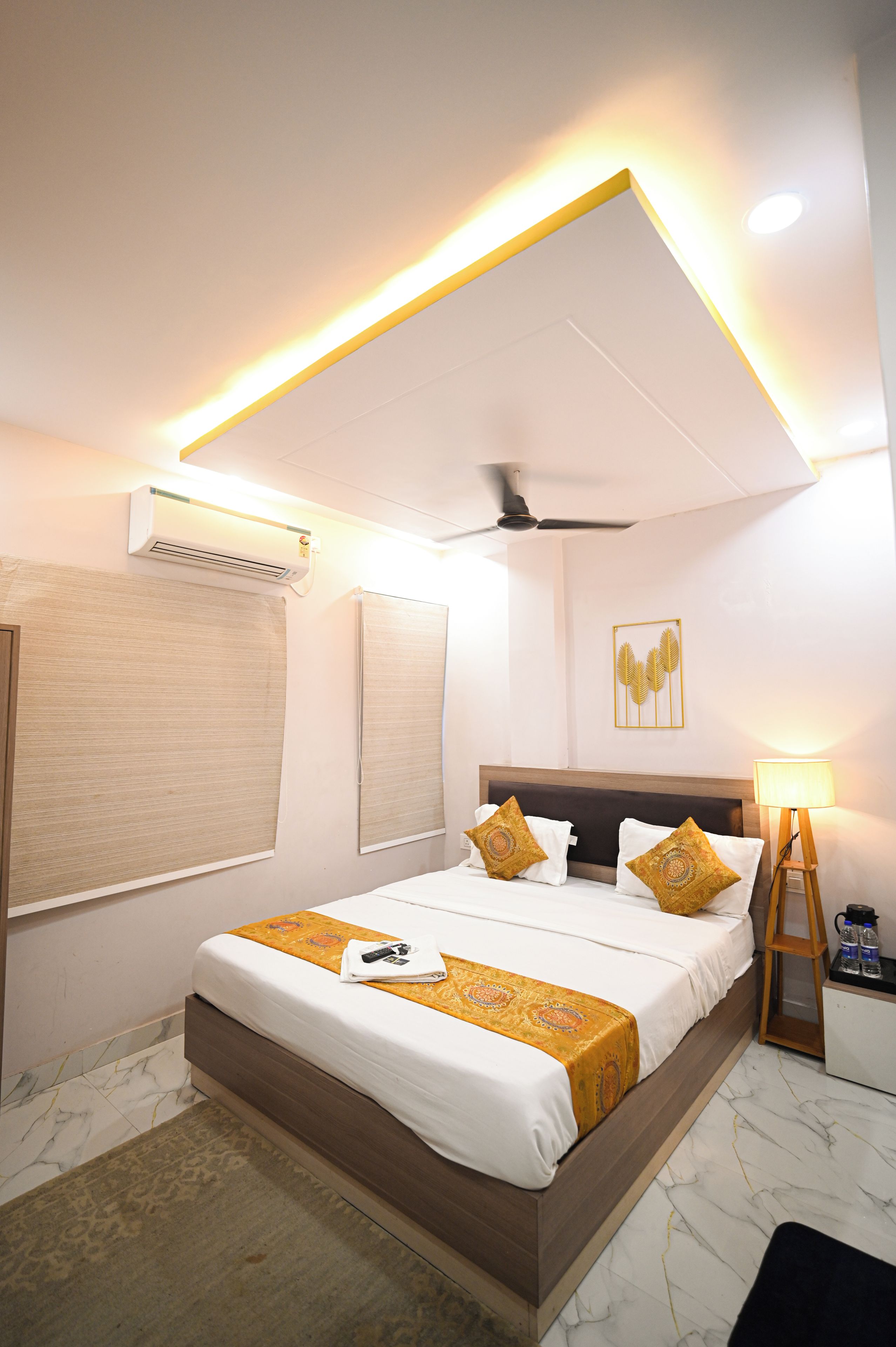 Quarto de hotel com cama, ventilador de teto, aparelho de ar condicionado, abajur e uma pequena mesa com garrafas.