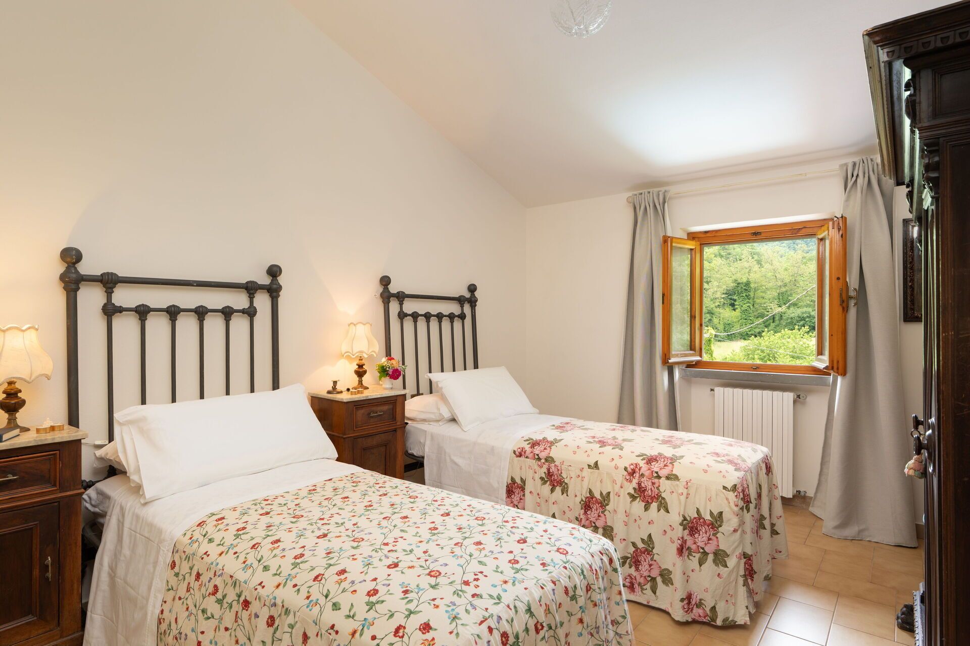 Een slaapkamer met twee enkele bedden, een nachtkastje met een lamp, een raam met uitzicht op groen en een houten garderobe.