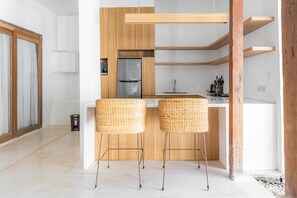 A modern kitchen with wooden cabinets, a stainless steel refrigerator, and two wicker bar stools. - Villa Elora Canggu (Canggu)