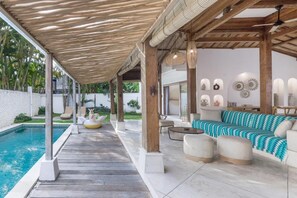 A covered outdoor area with a pool, wooden pillars, and a seating area with a patterned sofa and ottomans. - Villa Elora Canggu (Canggu)