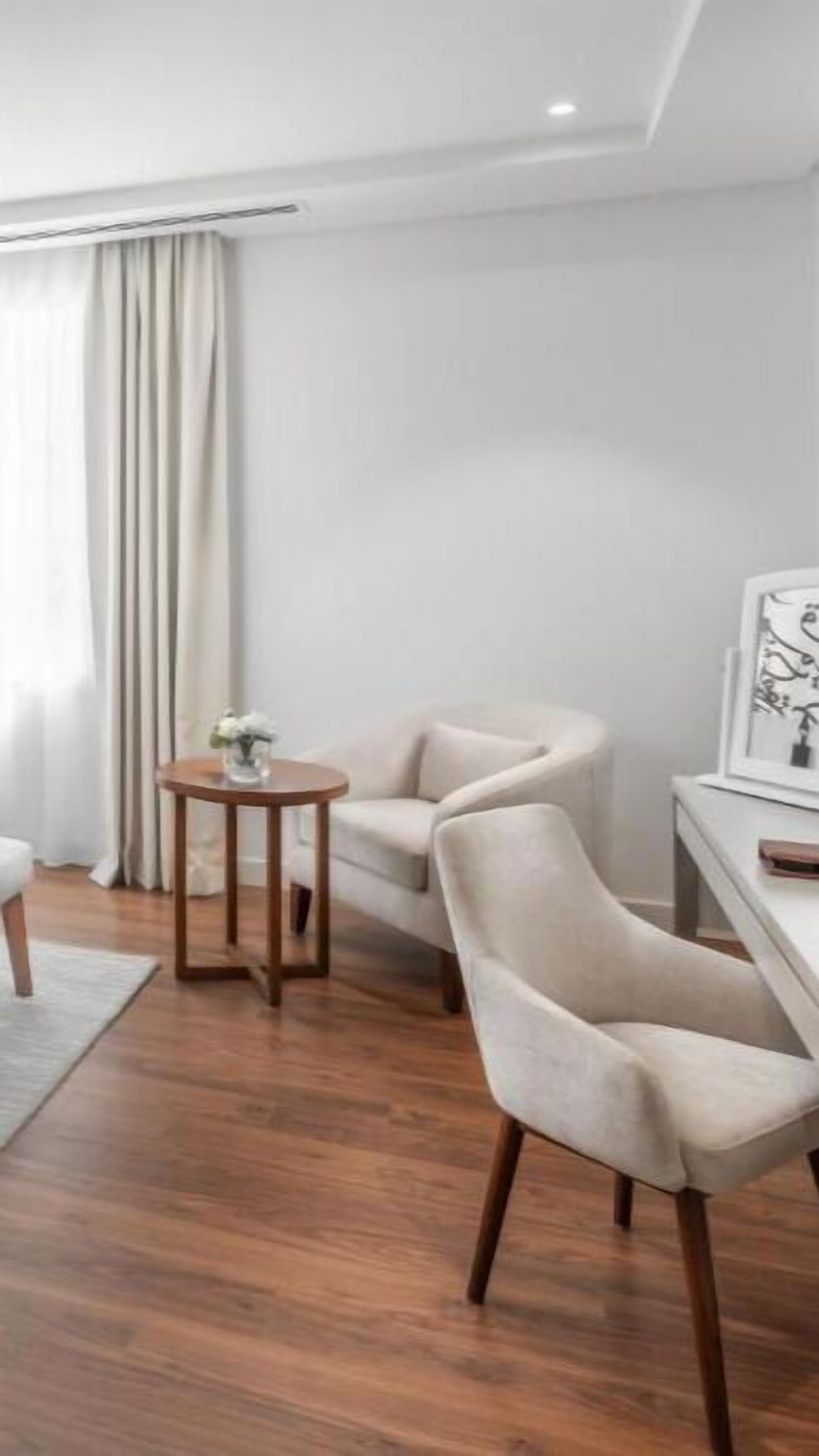 A modern, minimalist room with a wooden floor, a round wooden table with a vase of flowers, a beige armchair, and a white desk with a framed picture.
