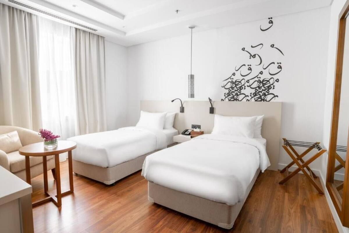 A hotel room with two beds, a small table with a vase, a sofa, and a wall art piece.