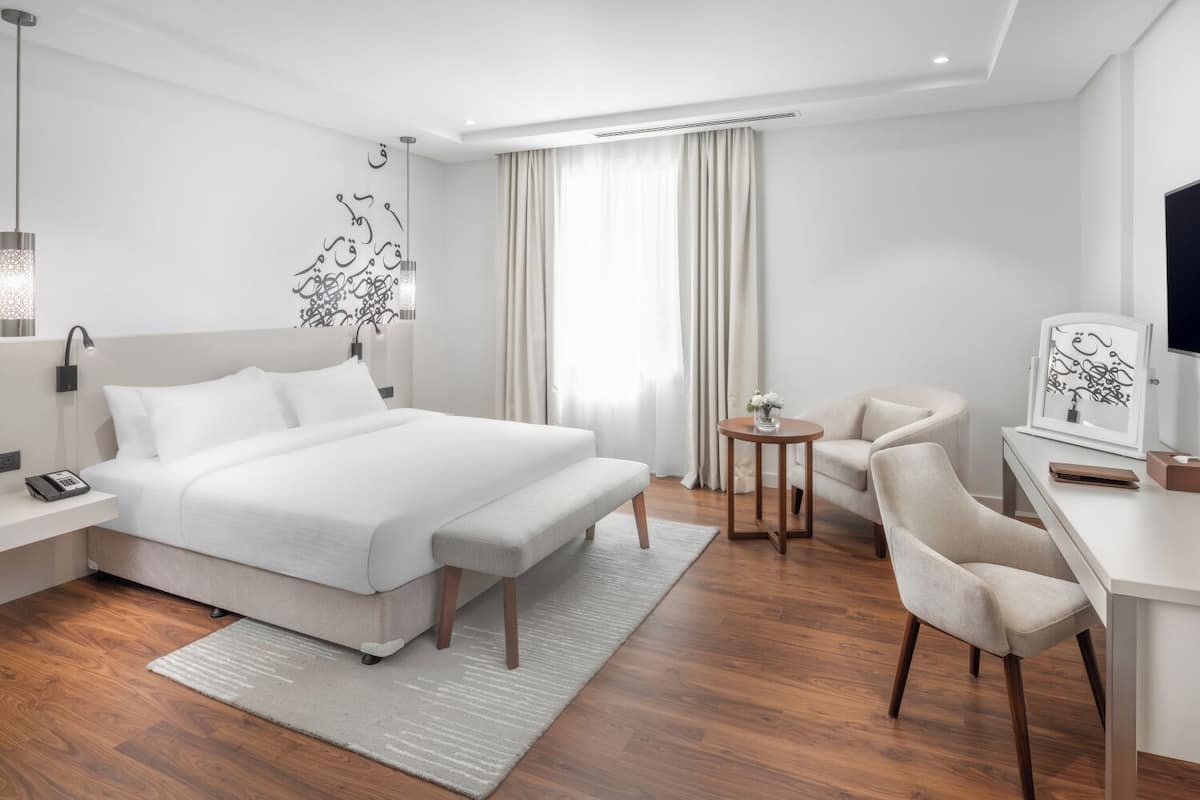 Quarto de hotel moderno com uma cama grande, mesa de cabeceira, abajur e uma escrivaninha com cadeira.