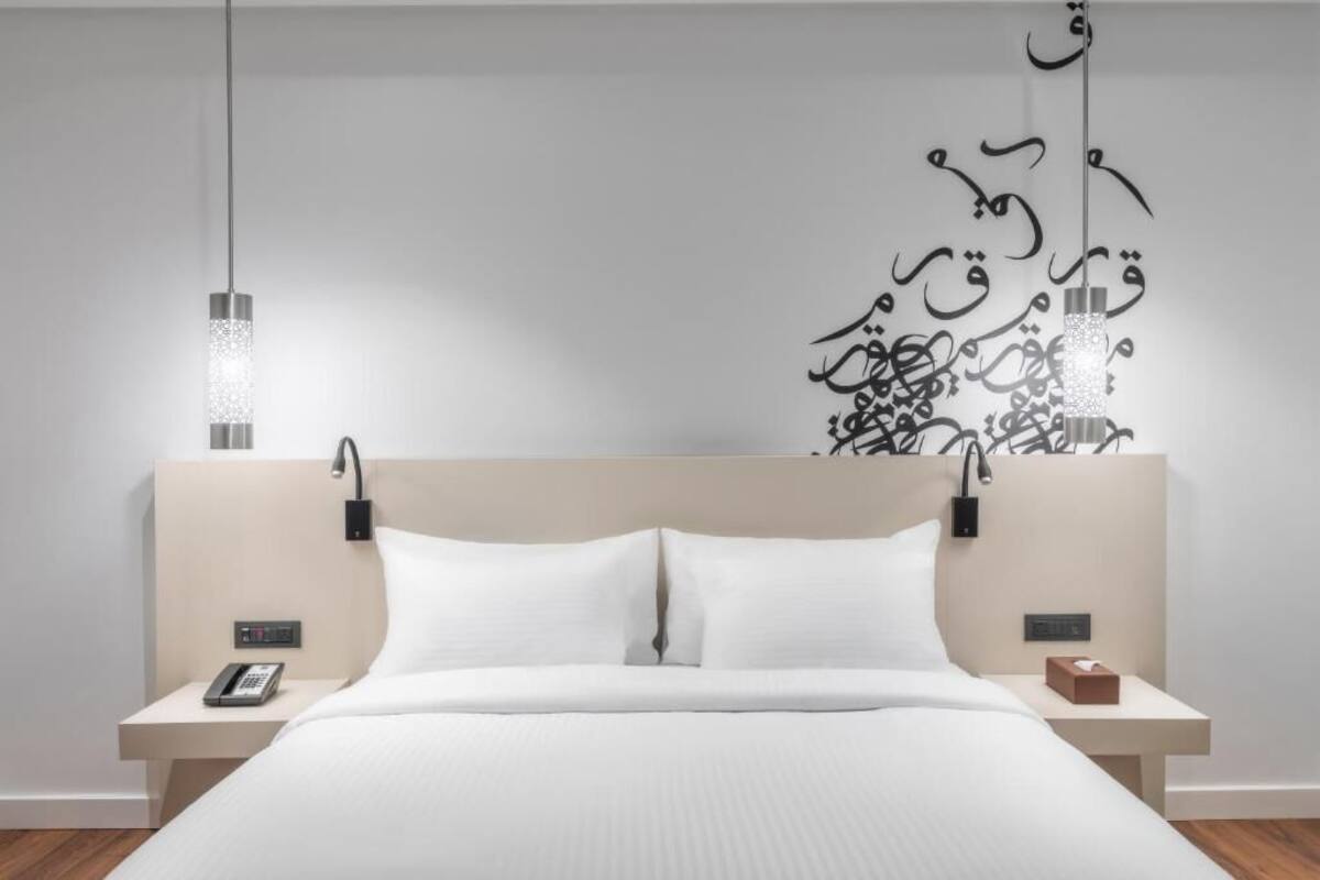 Quarto de hotel moderno com cama, mesas de cabeceira, telefone e um objeto decorativo na parede.