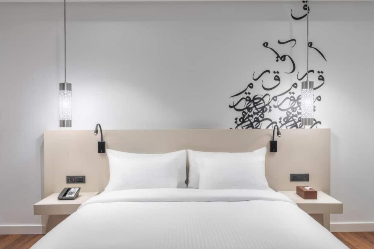 Quarto de hotel moderno com cama, mesas de cabeceira, telefone e um objeto decorativo na parede.