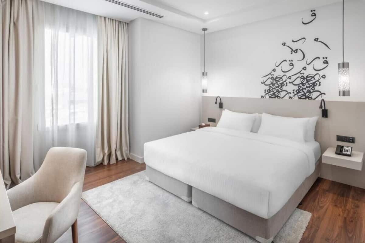 A modern hotel room with a large bed, a chair, a nightstand, and a wall with Arabic calligraphy.