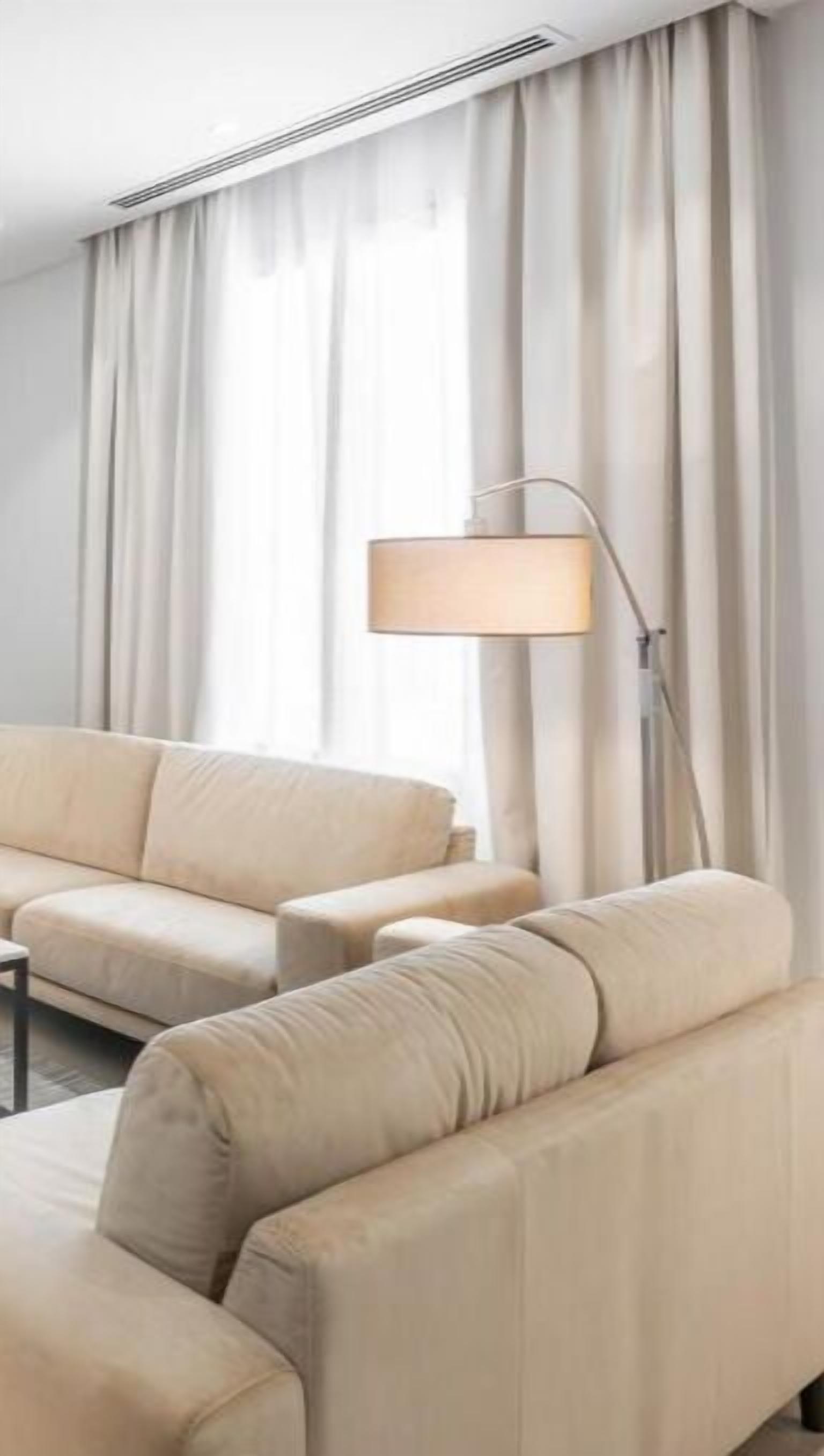 Una habitación de hotel moderna con un sofá beige, una lámpara de pie y cortinas de gasa.