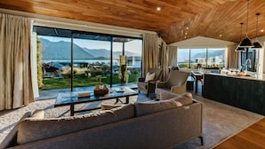 Un salon moderne avec un grand canapé, une table basse et un coin repas doté d’un bar. De grandes fenêtres offrent une vue sur un lac et des montagnes.