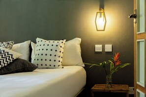 A neatly made bed with patterned pillows, a wall-mounted light fixture, and a small wooden nightstand with a plant.