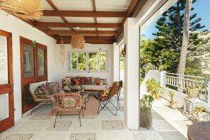 A covered outdoor seating area with a sofa, chairs, and a table, surrounded by potted plants and a view of trees.