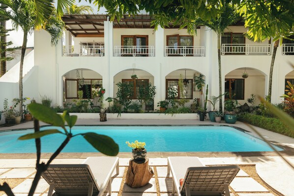 A poolside area with lounge chairs, a wooden stump with a flower pot, and a building with multiple balconies and windows.