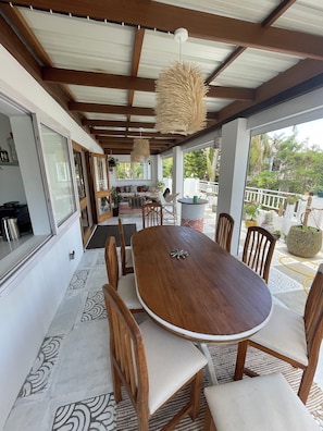 A covered outdoor dining area with a wooden table and chairs, a hanging light fixture, and a view of the outdoors.