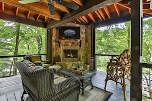 A covered outdoor seating area with wicker furniture, a stone fireplace, and a television mounted on the wall.