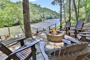 A riverside seating area with a fire pit, wooden chairs, and a stone-paved deck. - Whisperbend Lodge | Ellijay, GA (Ellijay)