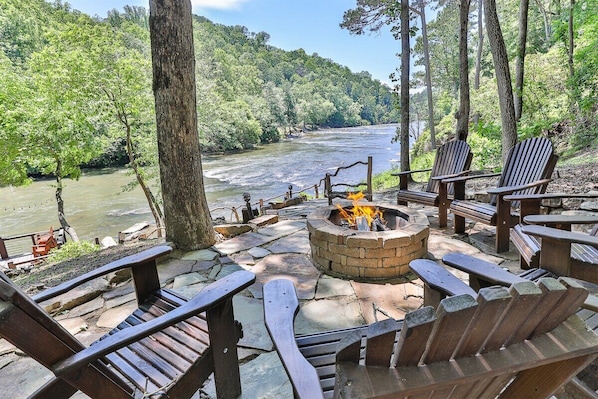 A riverside seating area with a fire pit, wooden chairs, and a stone-paved deck.